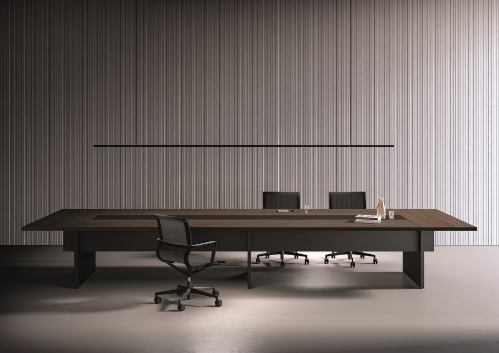 Un lungo tavolo rettangolare da sala riunioni, di colore scuro, con tre sedie da ufficio nere, addossato a una parete a coste grigio chiaro.