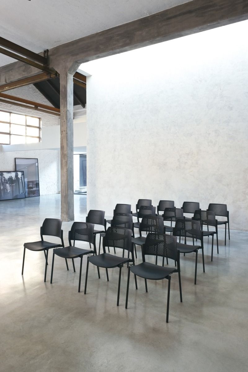 Un piccolo gruppo di sedie nere disposte in file all'interno di un'ampia sala minimalista con pavimento in cemento e pareti bianche.