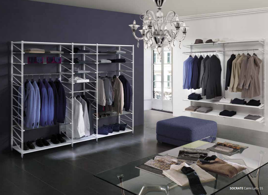 Una cabina armadio con scaffalature in filo metallico bianco piene di abiti appesi e piegati, un pouf centrale e un lampadario.