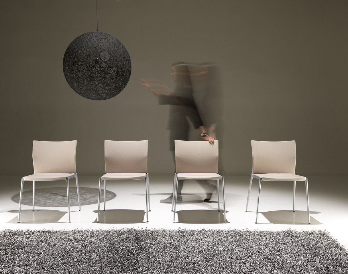 Quattro sedie beige allineate contro un muro, con una persona sfocata che si muove dietro la terza sedia, sotto una lampada rotonda.