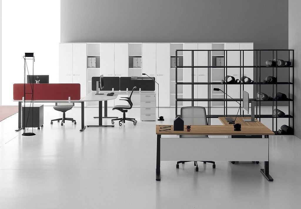 Un ufficio moderno con due scrivanie, una con un pannello divisorio rosso, e una scaffalatura in metallo nero addossata a una parete bianca.