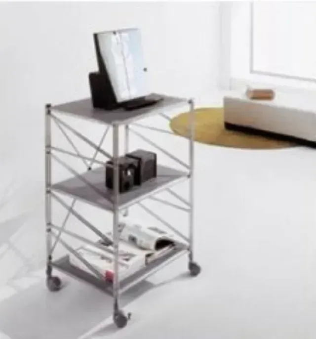 In una moderna stanza bianca, un carrello argentato a tre ripiani con ruote, dotato di mensole per apparecchi elettronici e libri.