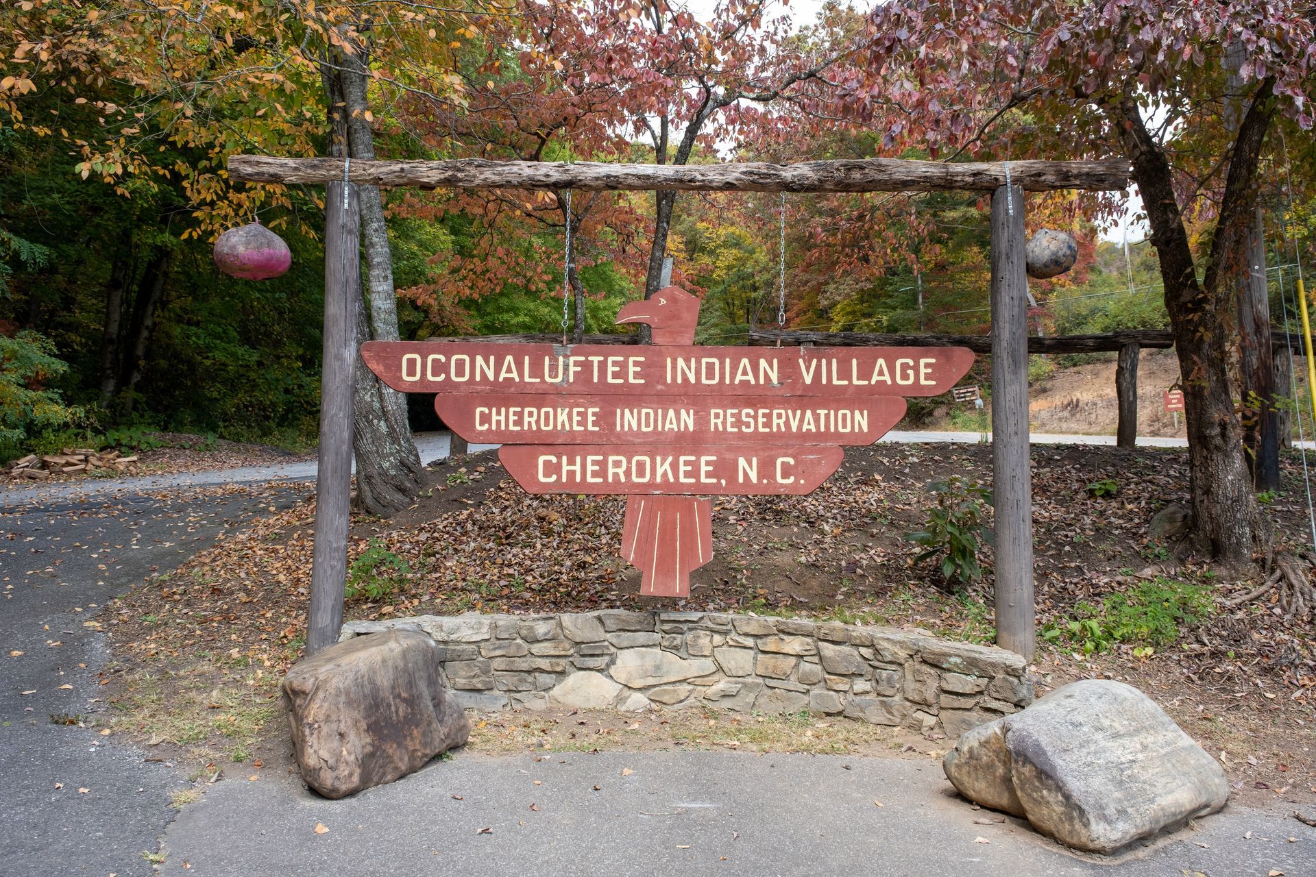 Sign for Oconaluftee Indian Village, Cherokee, N.C. in front of a road, with a red wooden sign and a stone base.