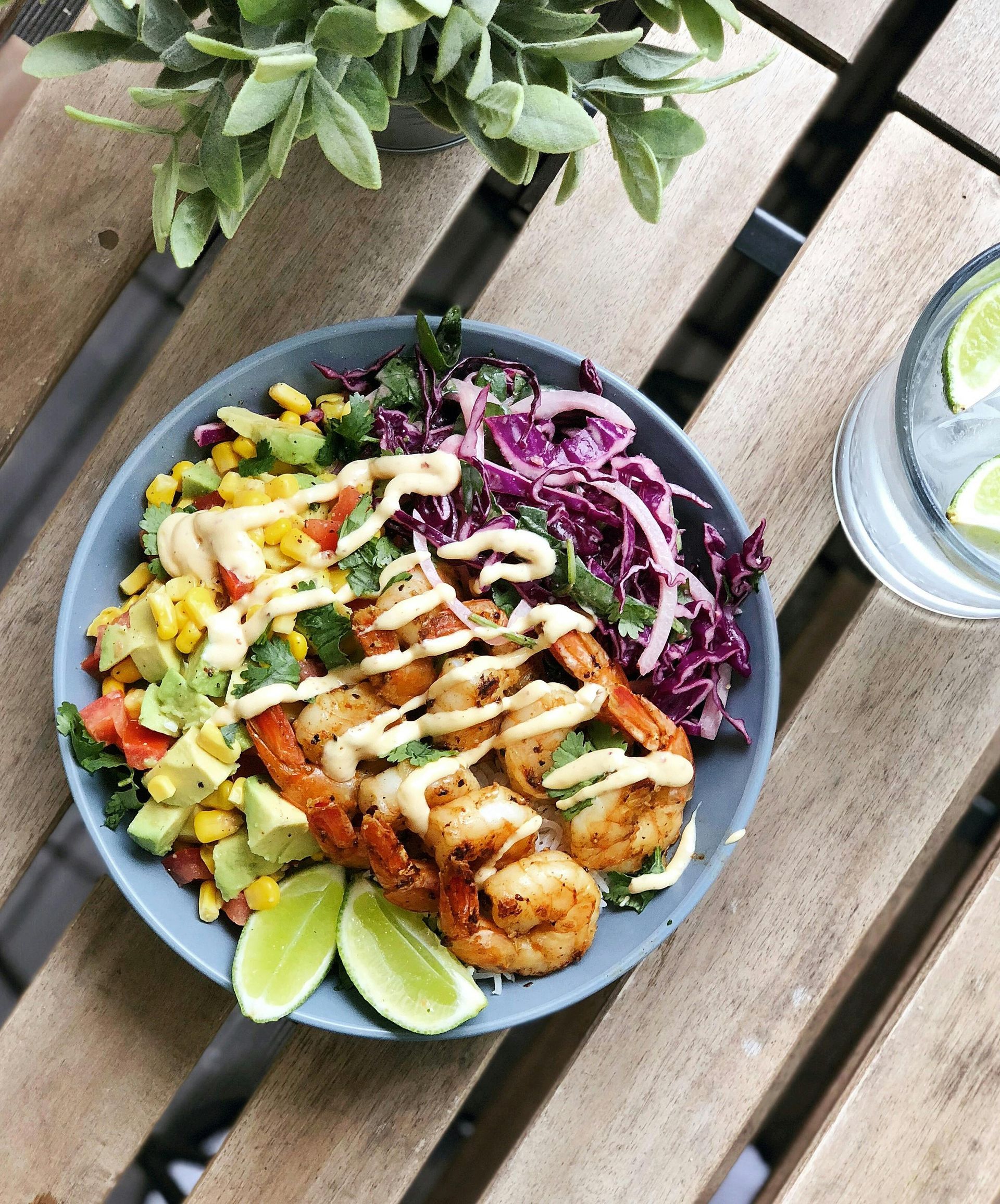 Shrimp bowl with avocado, corn, red cabbage, and dressing on a wooden table, with lime slices.
