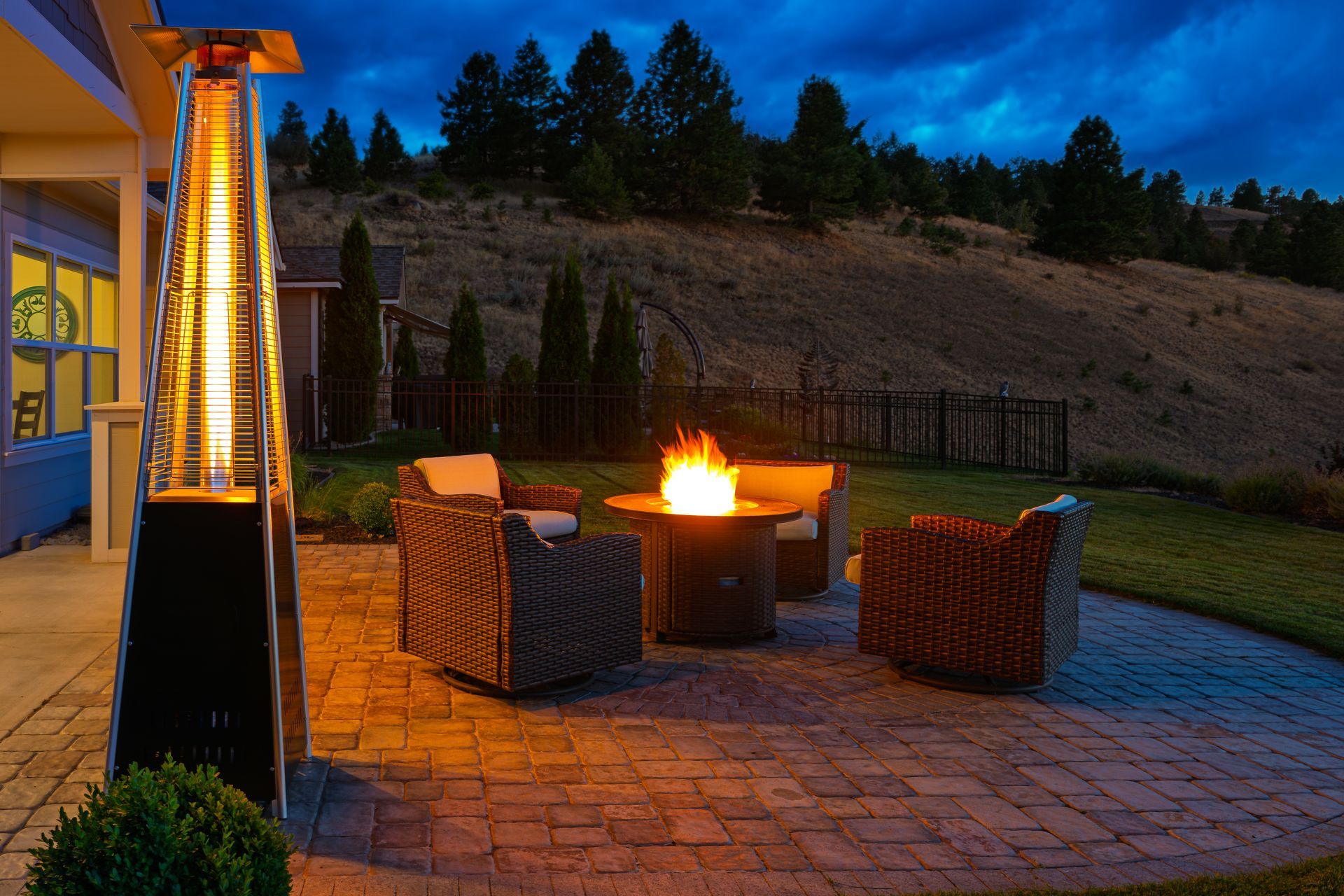 A patio at twilight featuring a tall, illuminated outdoor heater, a glowing fire pit, and three wicker chairs.