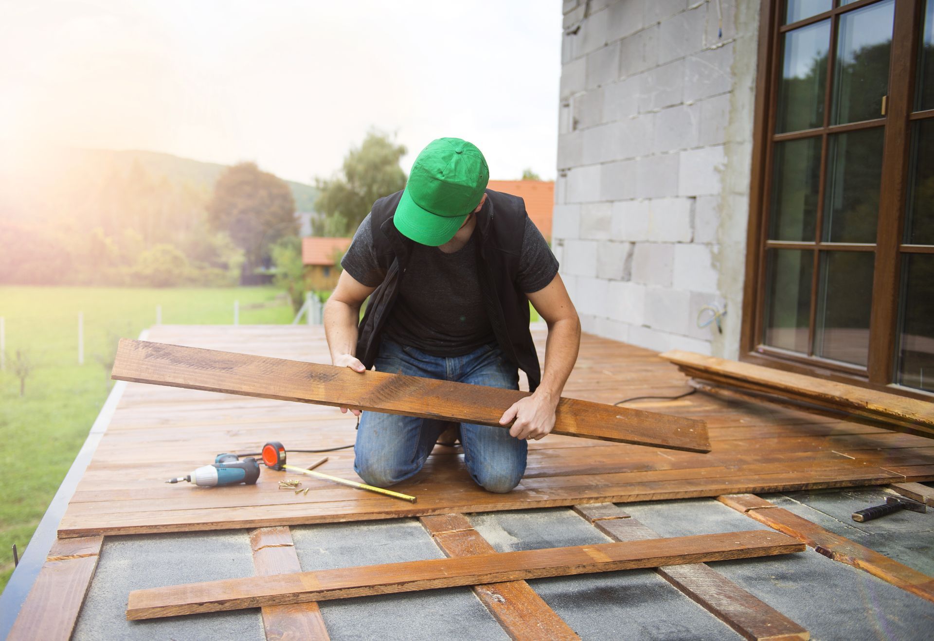 A person wearing a green cap and vest kneels on a wooden deck, measuring a floor plank for installation.