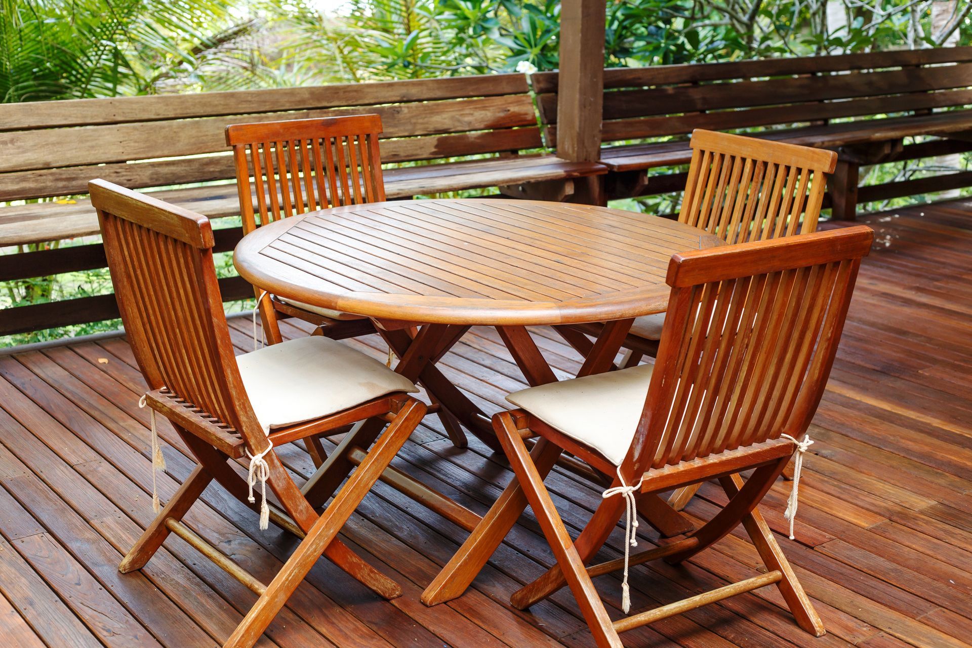 A round wooden patio table with four folding chairs featuring white cushions, set on a wooden deck.