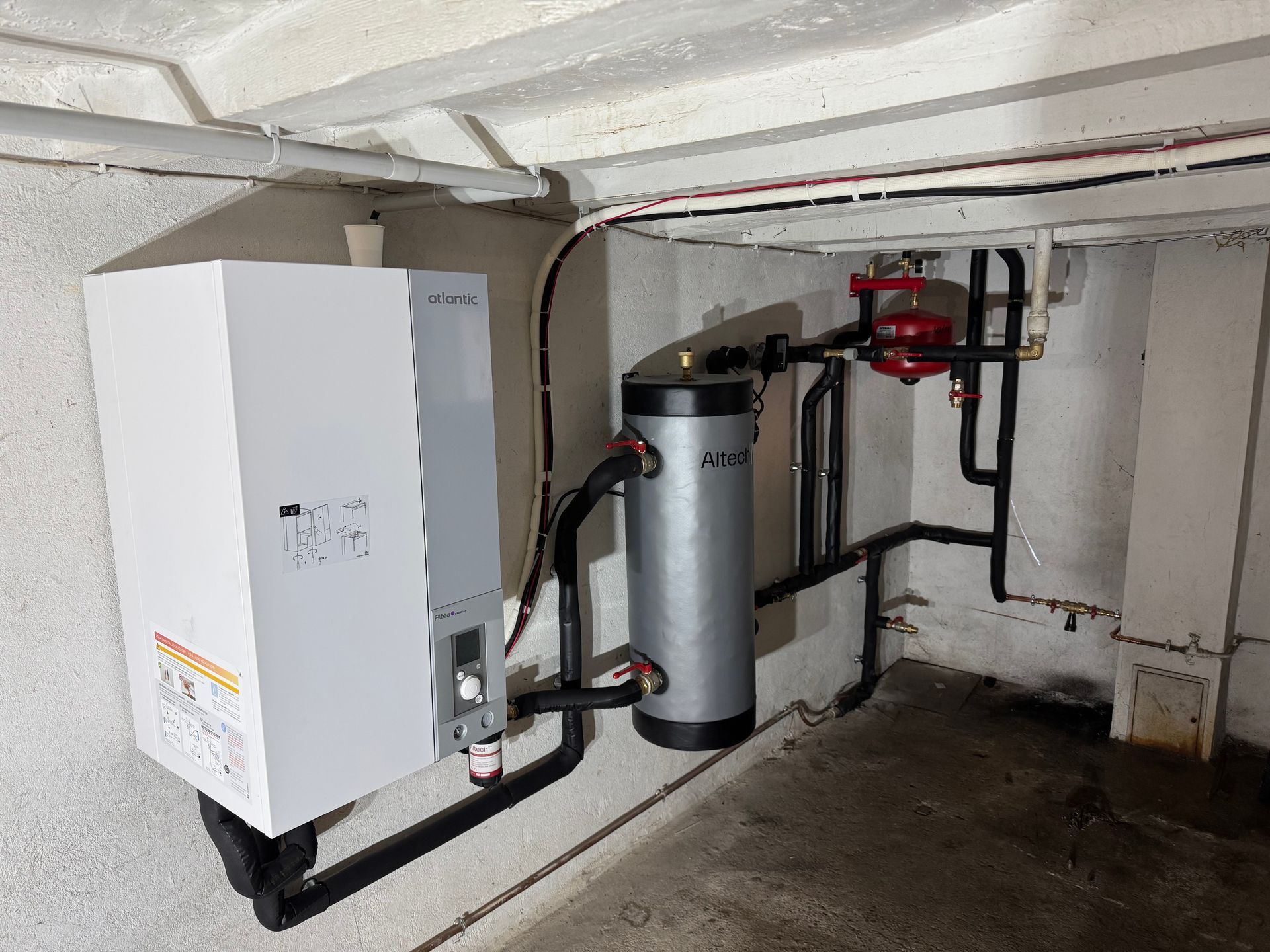 Chauffe-eau blanc et réservoir de stockage avec tuyaux noirs contre un mur en béton.