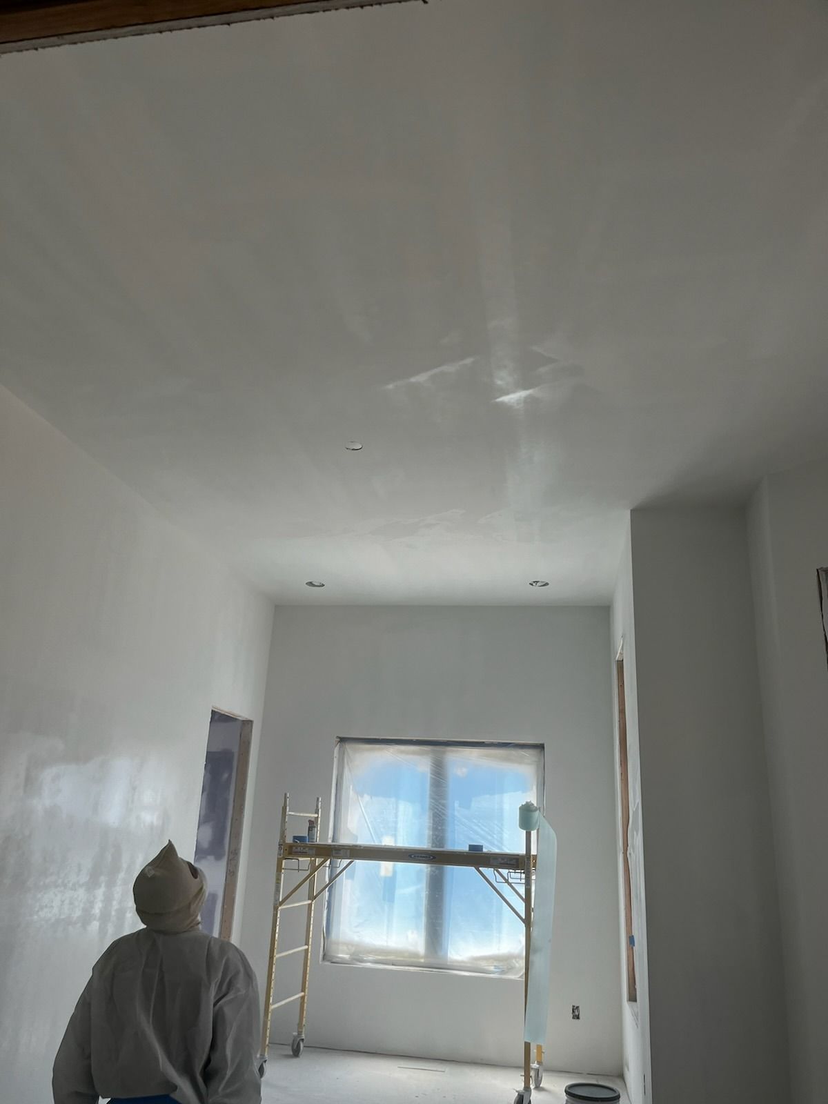 Person looking at a freshly plastered white ceiling in a room with a window and scaffolding.