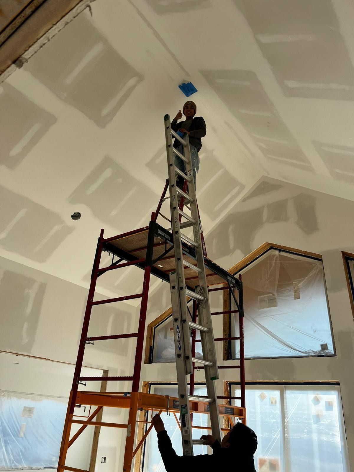 Construction worker on tall ladder reaching towards the ceiling, another person below. Interior.