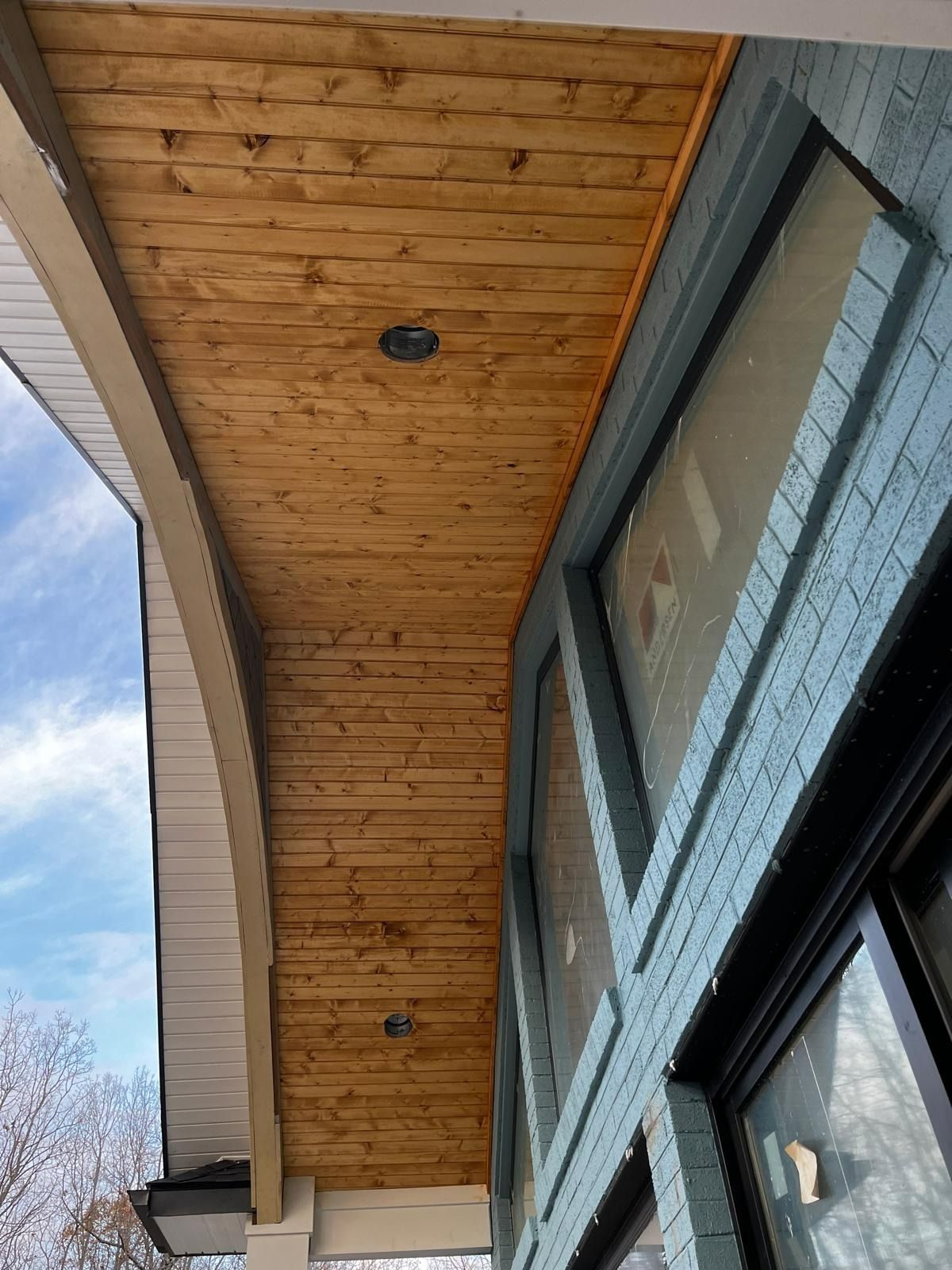 Wooden porch ceiling with recessed lighting next to a building with a blue brick exterior.
