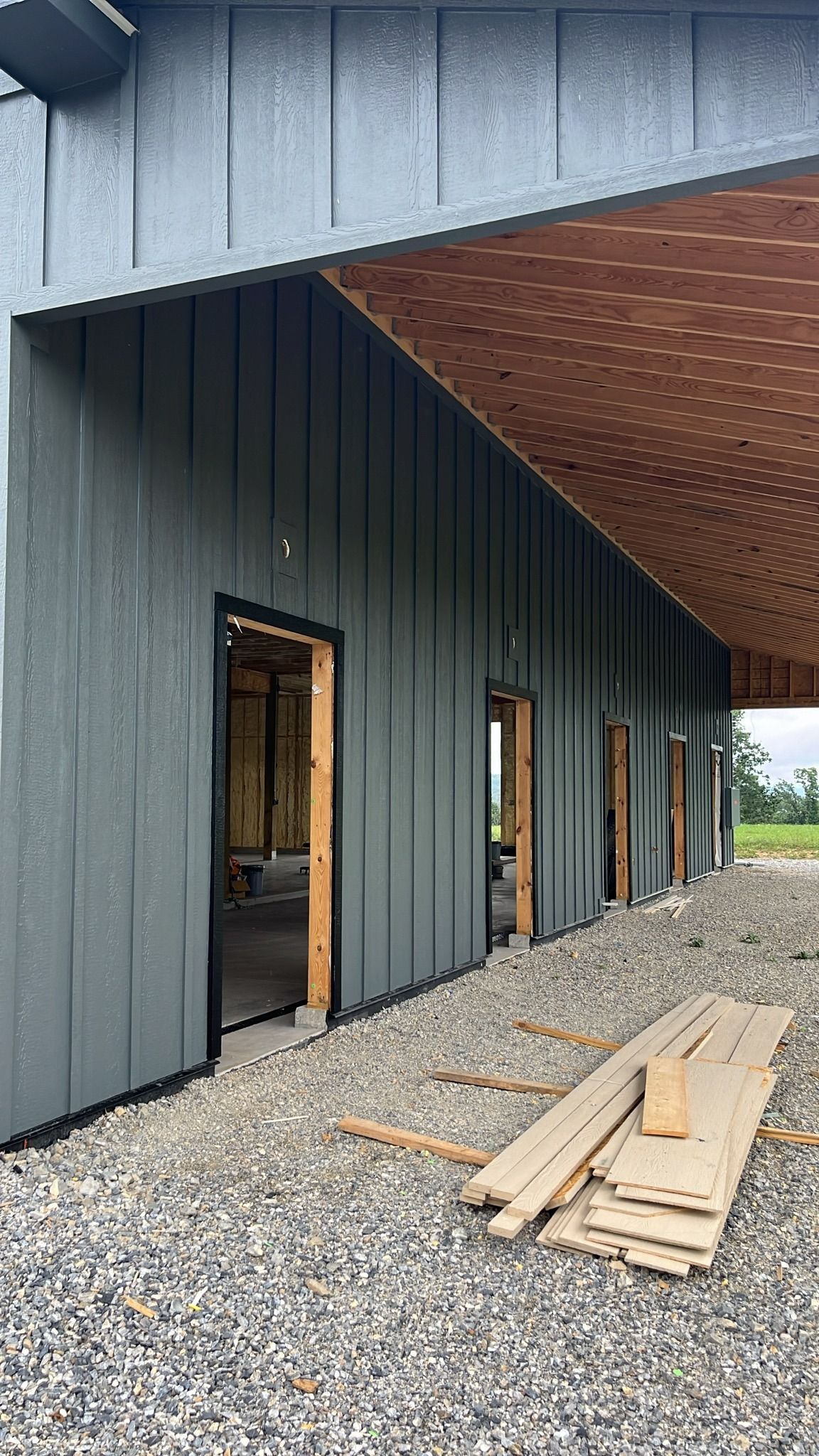 Building exterior with dark gray siding, open doorways, and gravel ground, under construction.