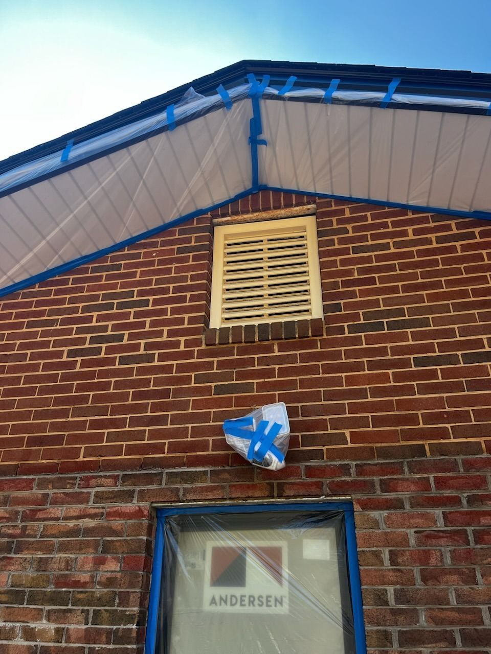 Brick building with blue-taped trim, a window, and a vent; plastic sheeting protects the facade during construction.