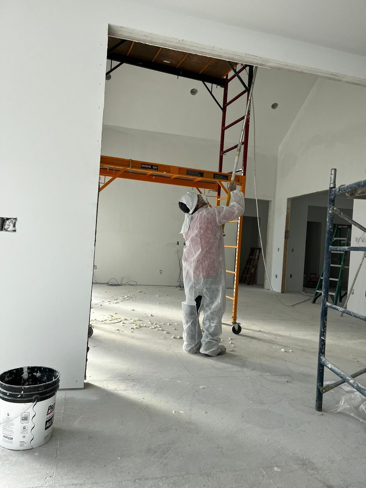 Person painting a room with a sprayer, wearing protective gear. Scaffolding, bucket, and unfinished walls visible.