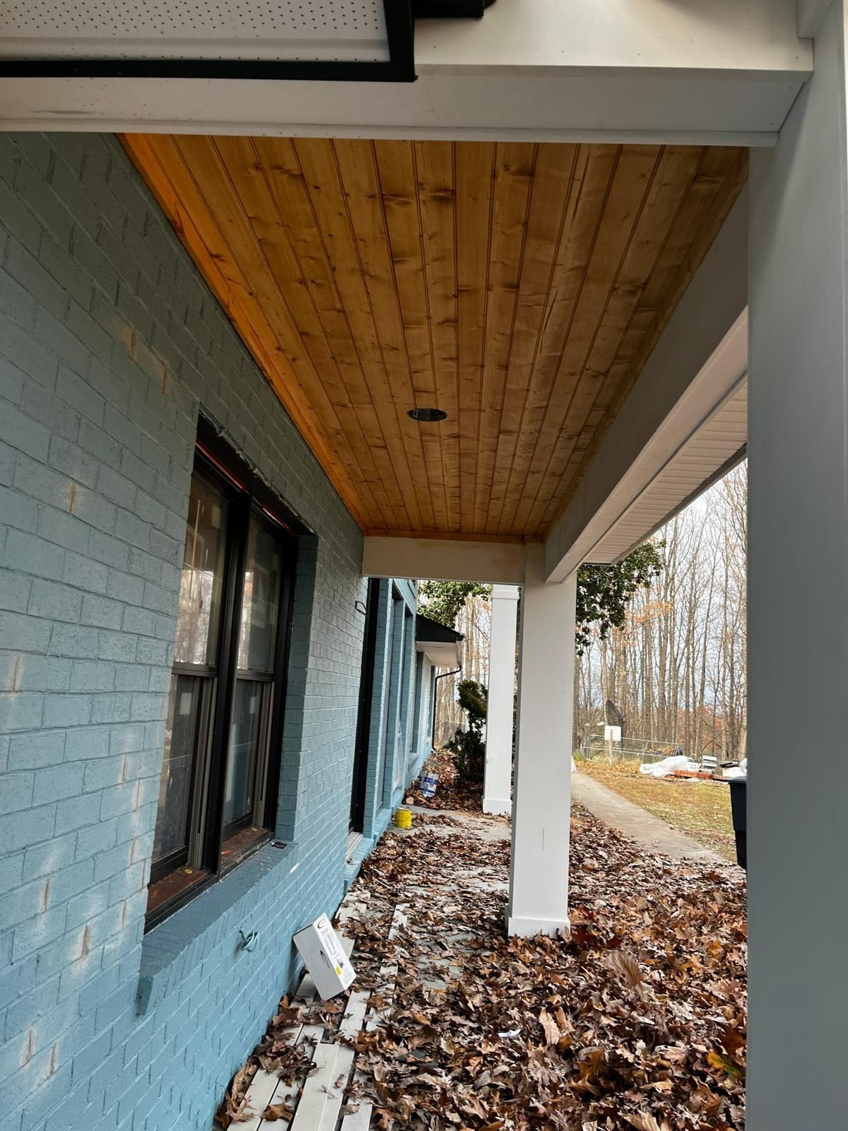 Blue brick house with a wooden porch ceiling, white supports, and fallen leaves on the ground.