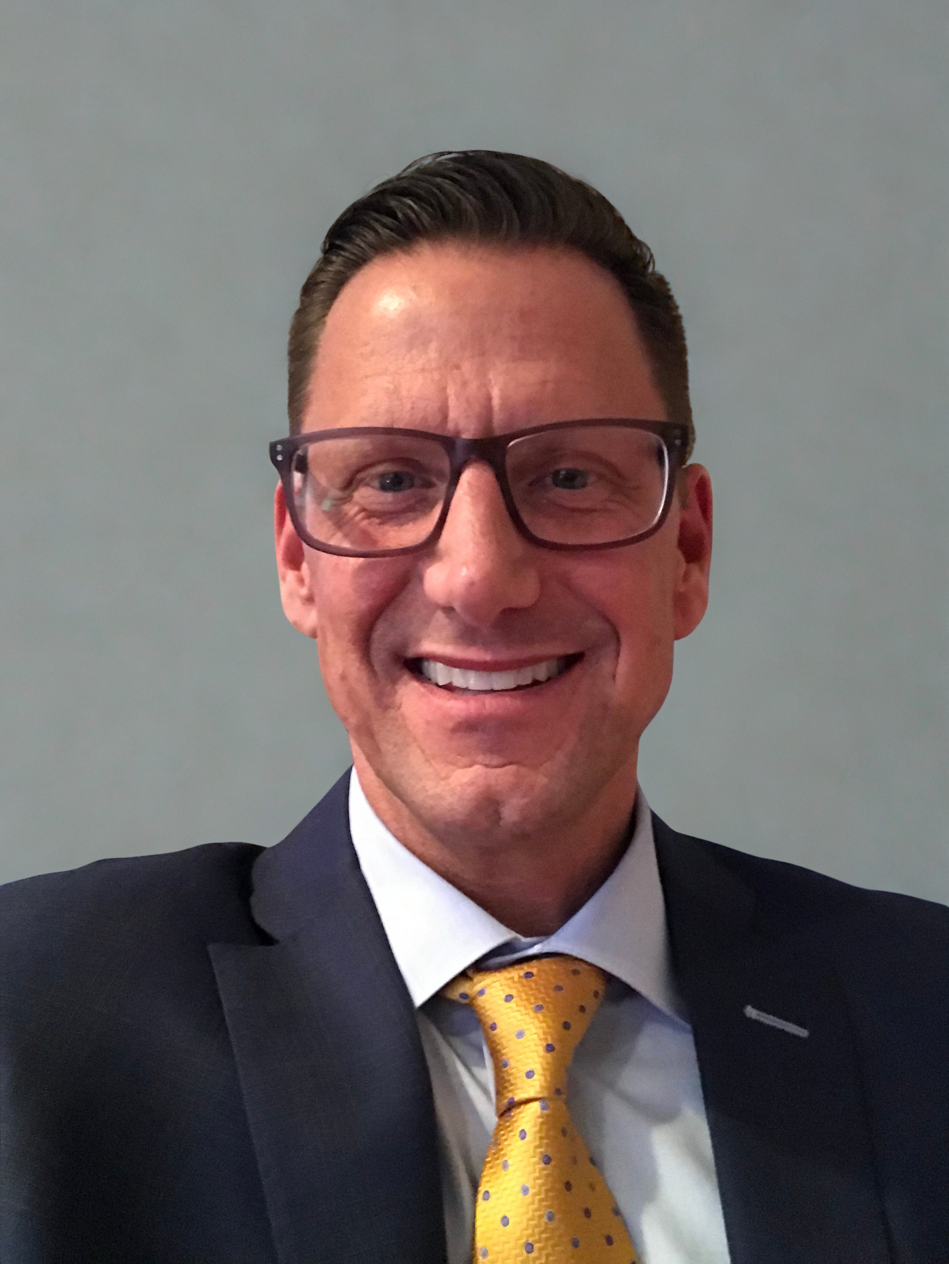 Man wearing glasses, dark suit, and yellow patterned tie smiling at the camera.