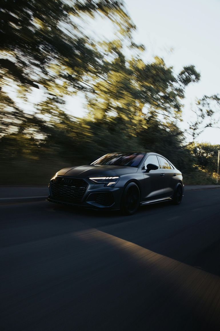 Dark gray car speeding on a road with blurred background of trees and sunlight.