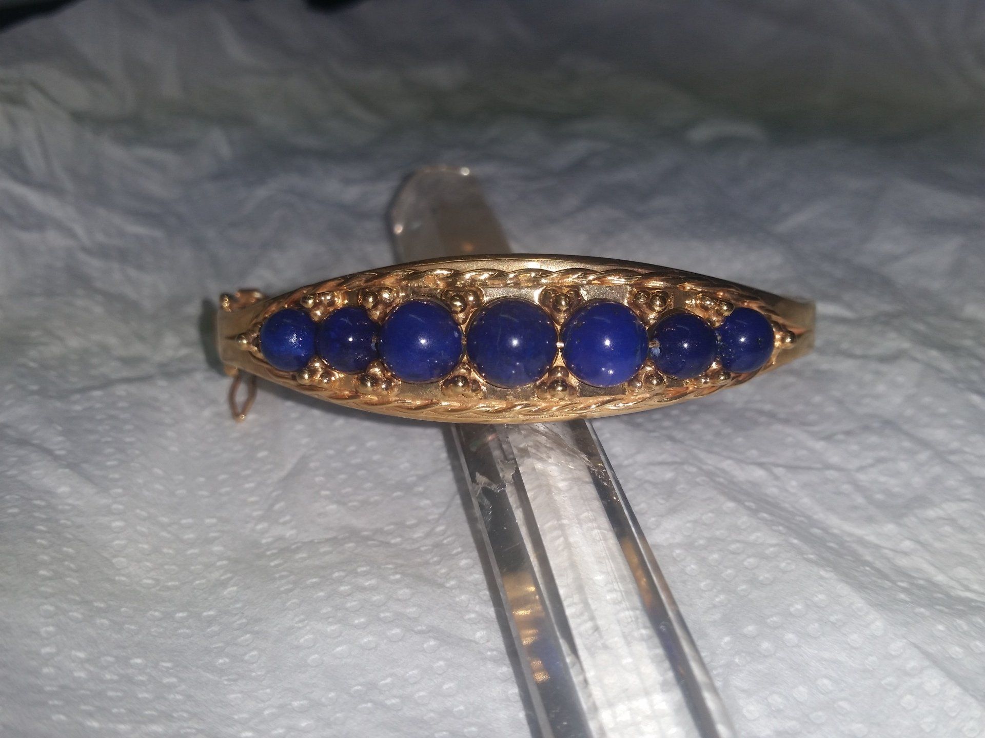 Un bracciale allungato color oro con sette perle rotonde di pietre preziose blu scuro incastonate su uno sfondo di metallo filigranato.