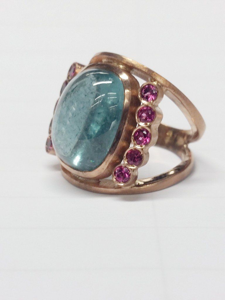 Anello in oro rosa con una grande gemma ovale color verde acqua affiancata da due file verticali di piccole pietre rotonde rosa.