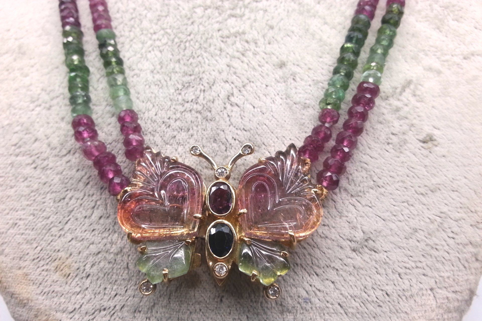 Un ciondolo a forma di farfalla con ali intagliate in pietre preziose multicolori, appeso a una collana a due fili di perline.