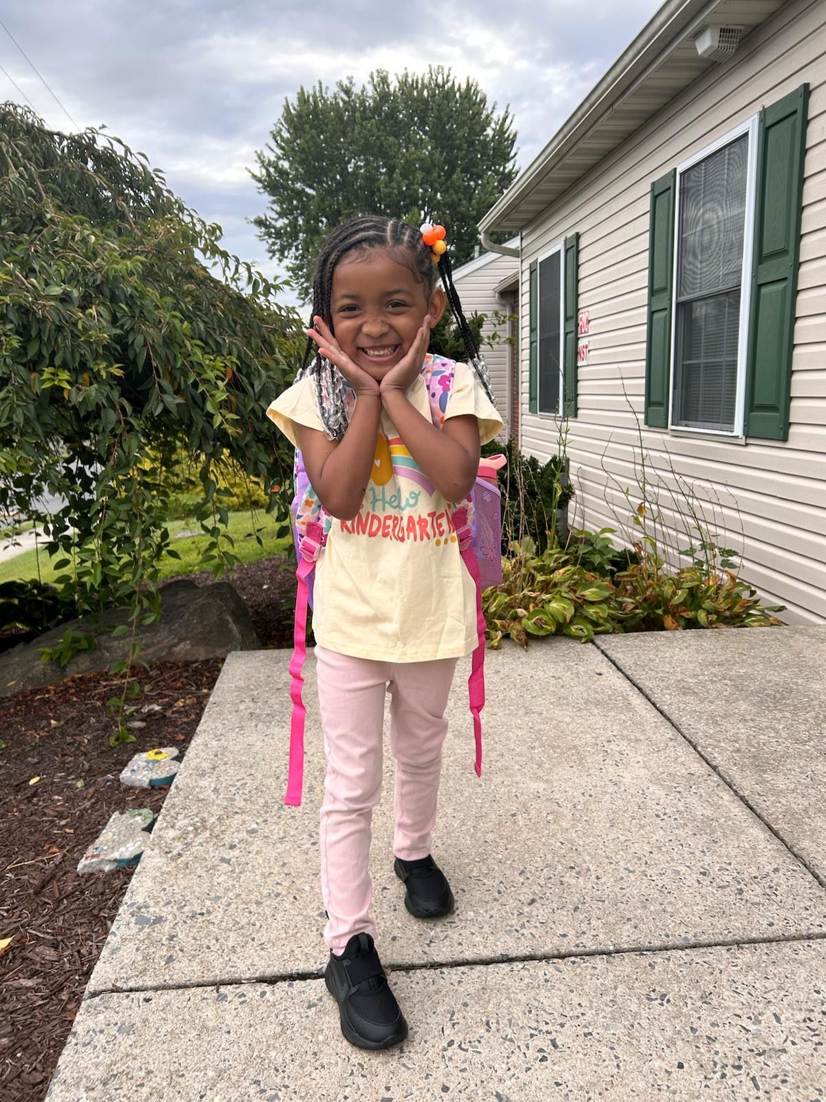 Girl with braided hair, pink pants, and backpack smiles, holding hands near her face, outside a house.