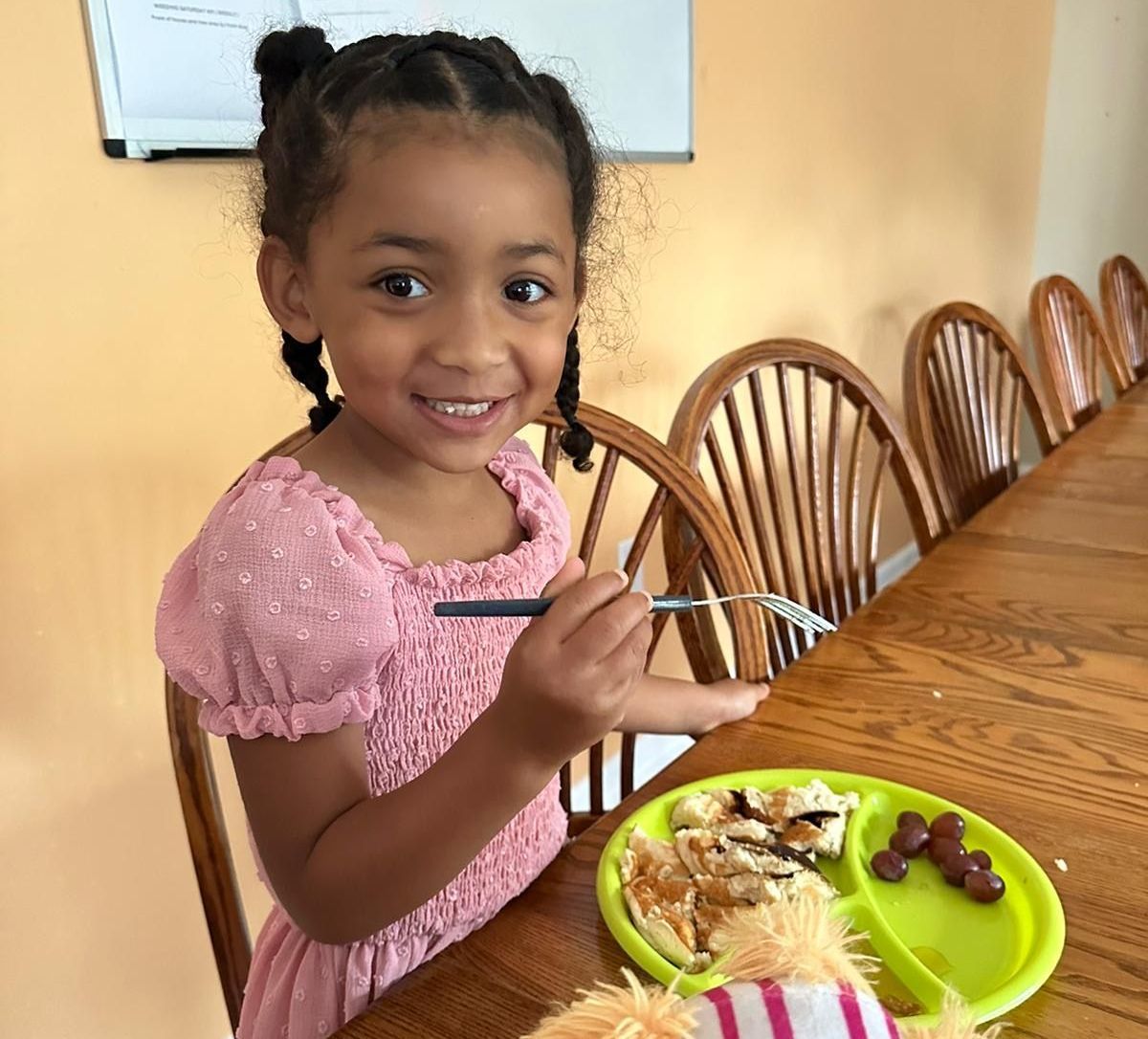 Young child smiles, eating from a green plate at a long wooden table.