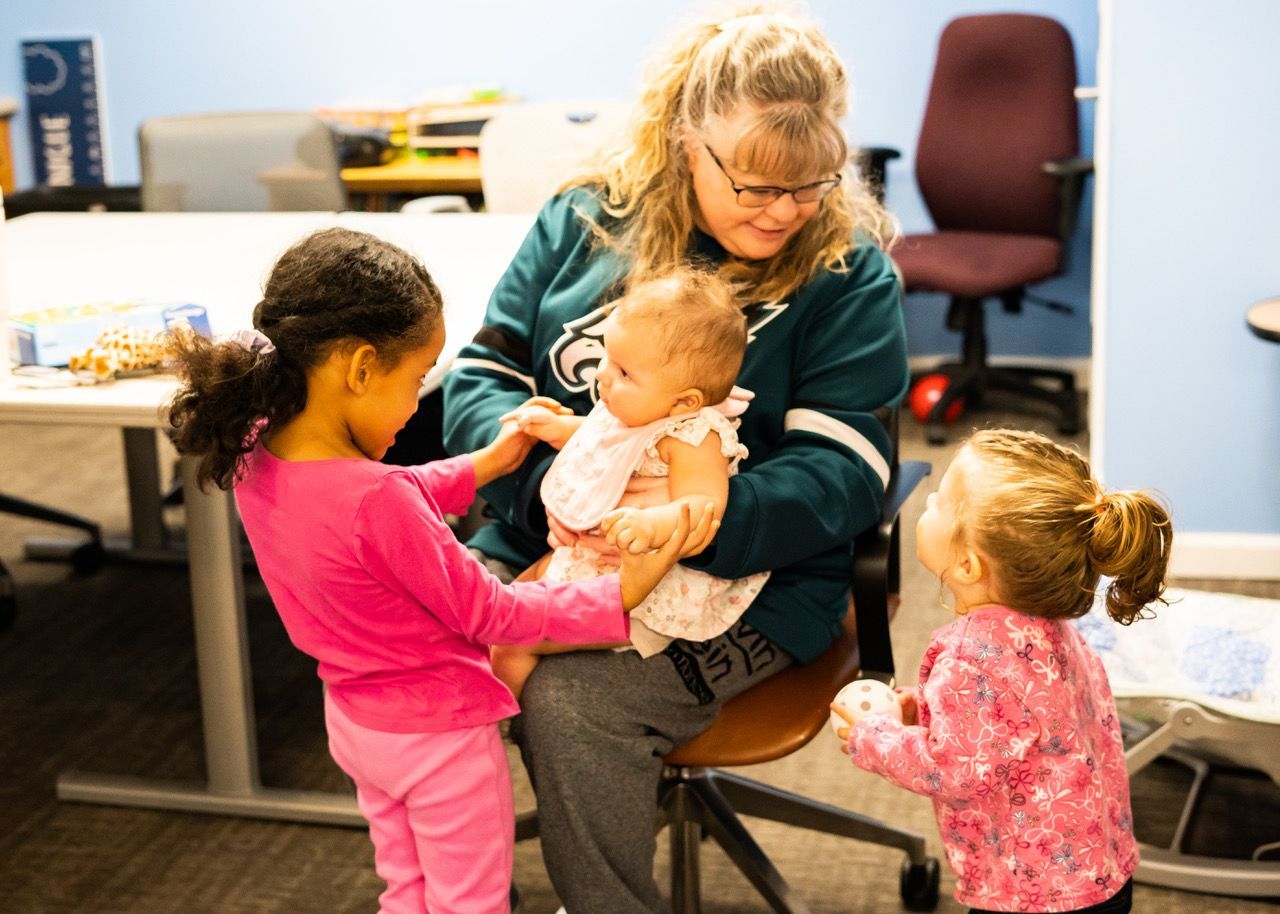 Woman holds baby, interacting with two young children in an office setting.