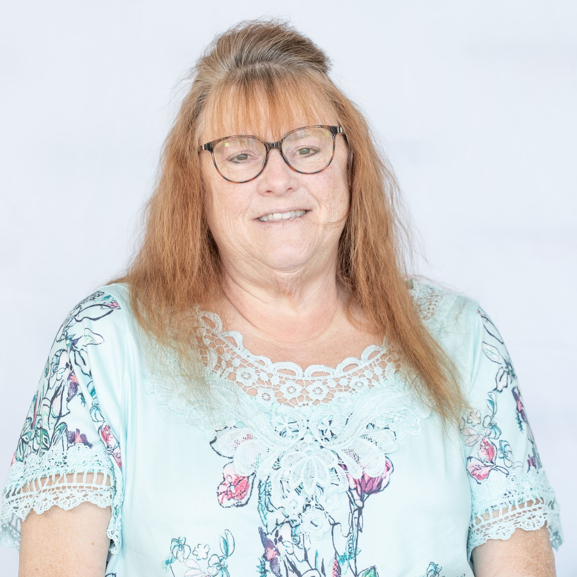 Woman with glasses, light blue top with floral design, smiling against a white backdrop.
