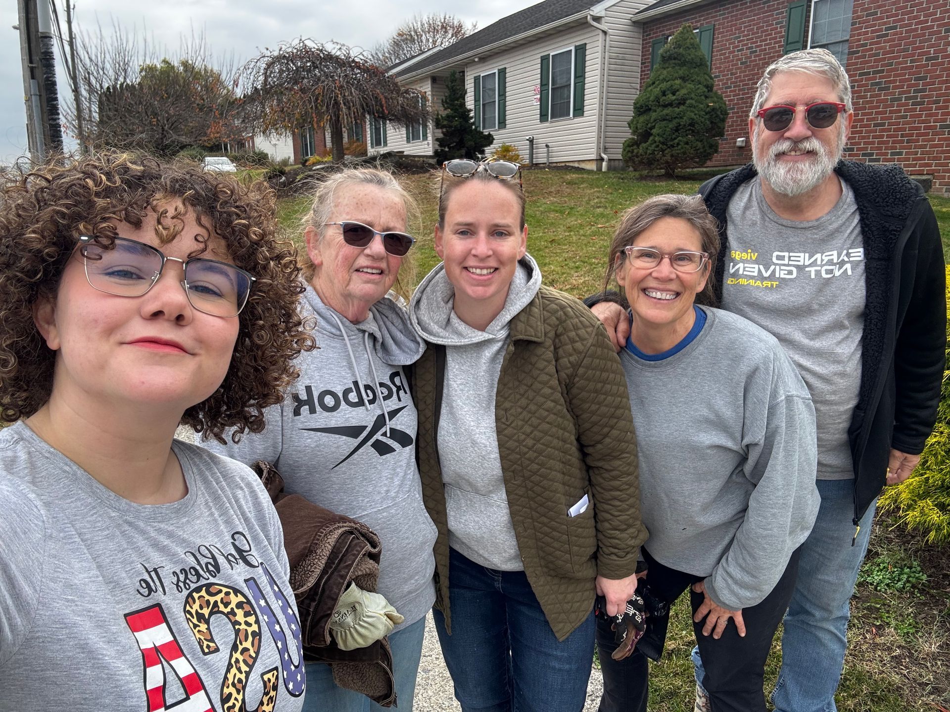 Five people smiling together outdoors, near a house. Overcast sky.