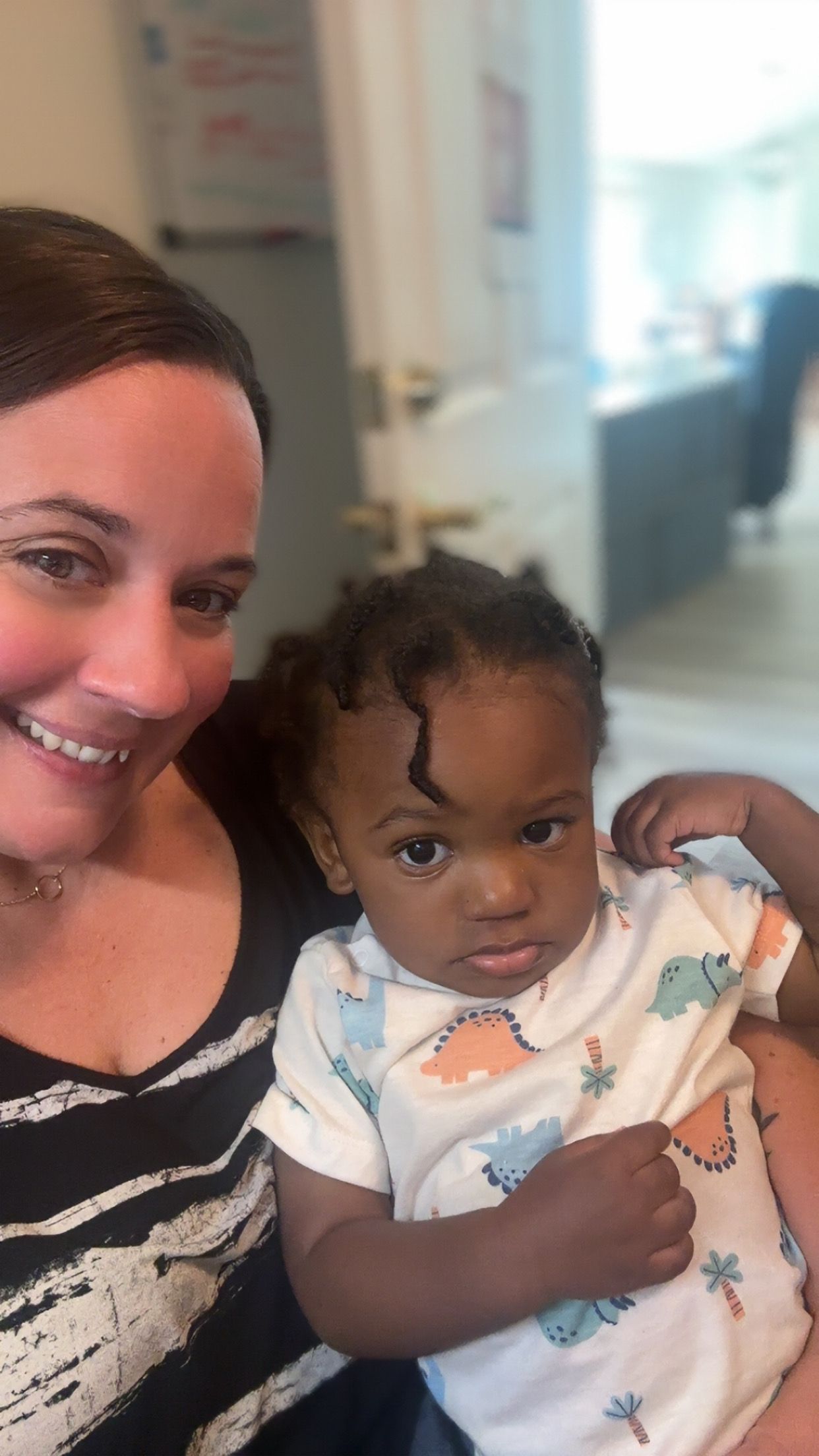 Woman smiling, holding a baby in a dinosaur print onesie. Interior setting.