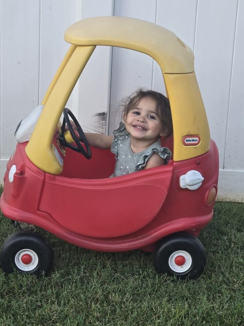 Child smiling in a red and yellow toy car outdoors.