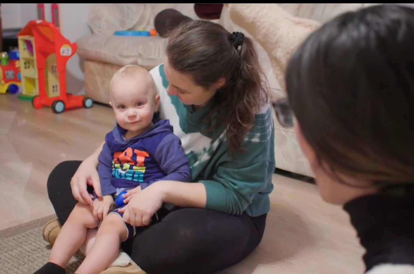 Woman reads a book with a baby on a gray couch in a living room, with a kitchen visible in the background.