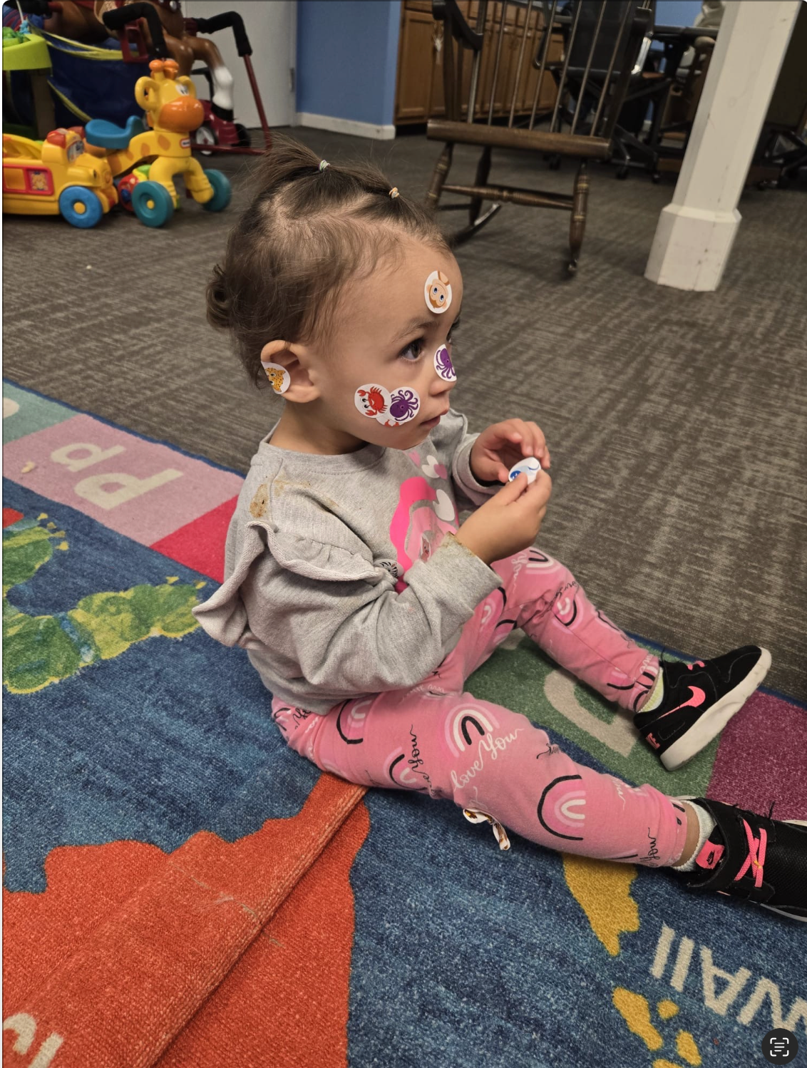 Child sitting on a colorful rug, wearing pink leggings and gray sweater, stickers on face.