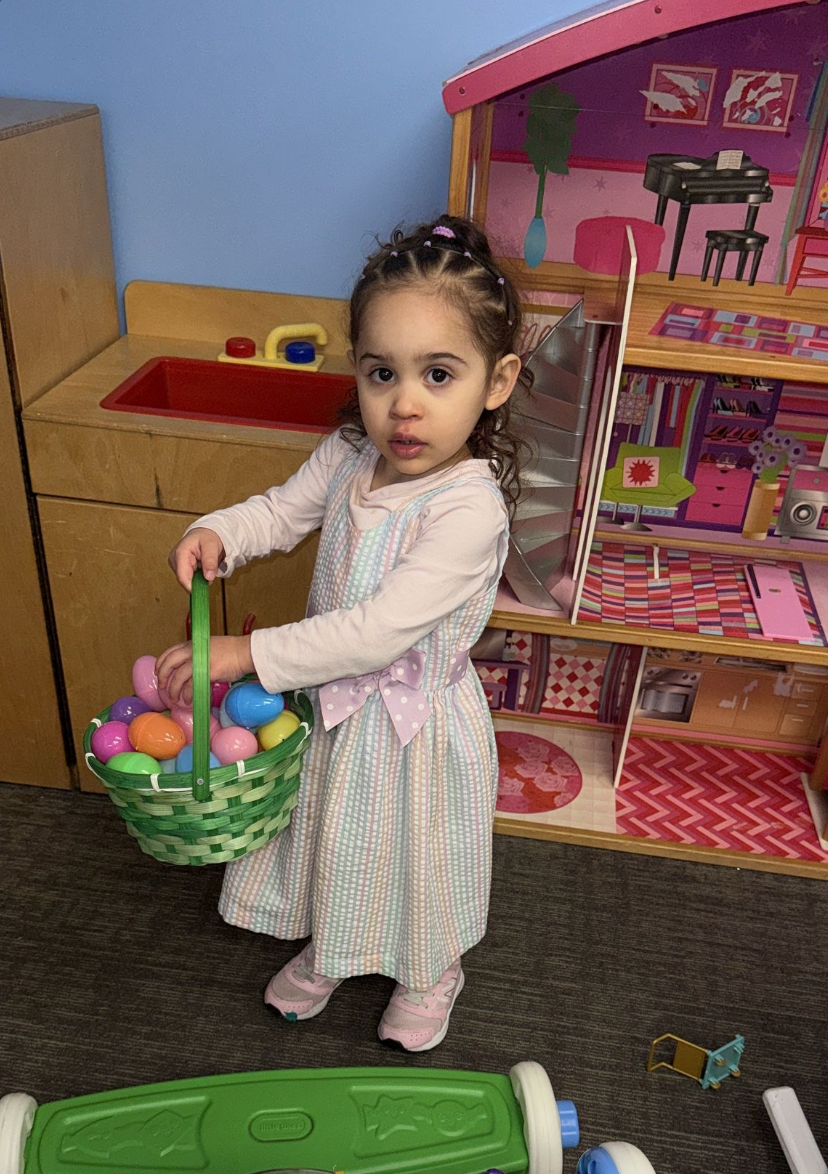Child holding a basket of Easter eggs, standing near a dollhouse and toy kitchen.