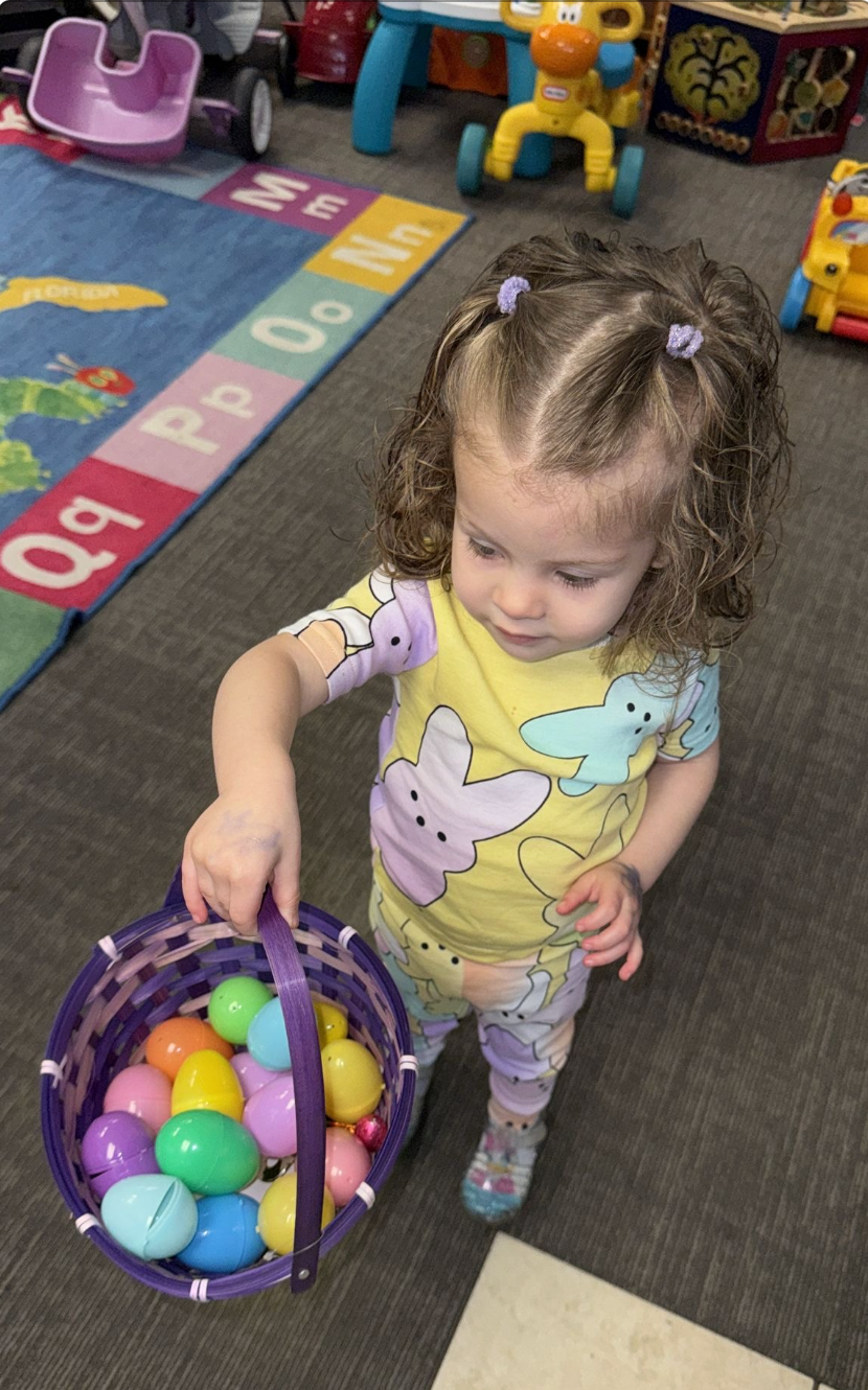 Child with curly hair holds a purple basket filled with colorful Easter eggs, indoors near a rug with letters.