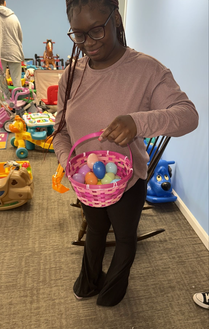 Person holding a pink Easter basket filled with colorful eggs, in a room with toys.