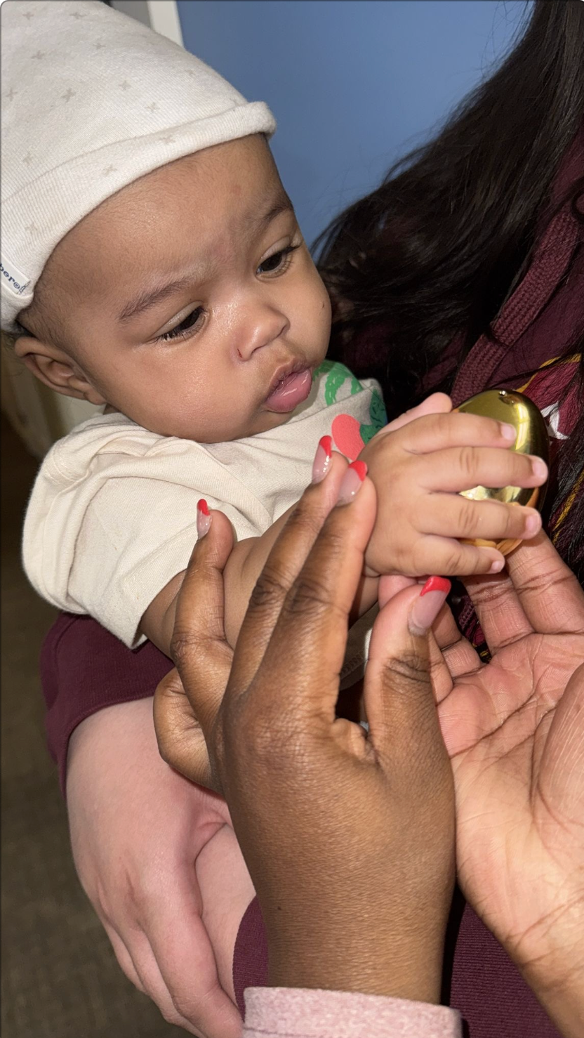 Baby holding a golden object, resting in someone's arms.  Person's hands with red nails interacting with the baby.