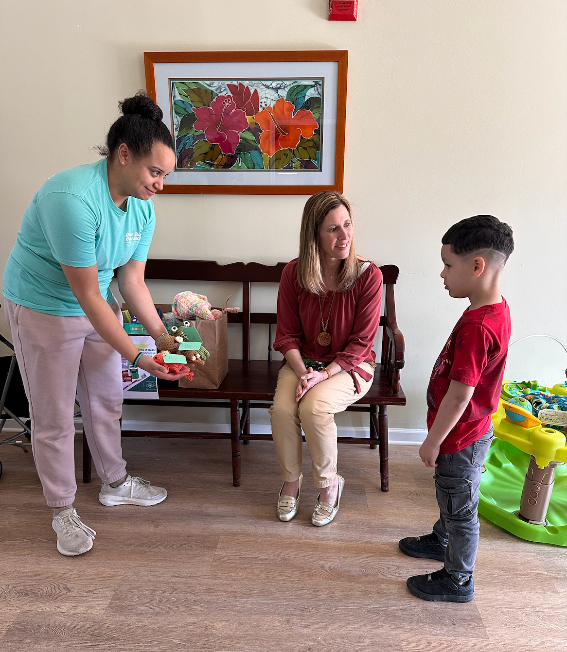 Woman offers gift to a child; another woman watches. Interior setting with bench and colorful painting.