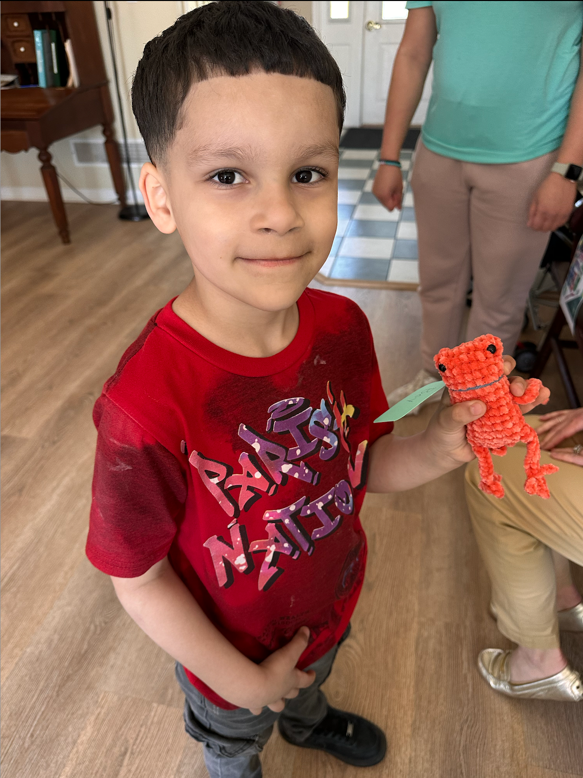 Boy holding an orange woven toy frog, smiling, wearing a red shirt with writing.
