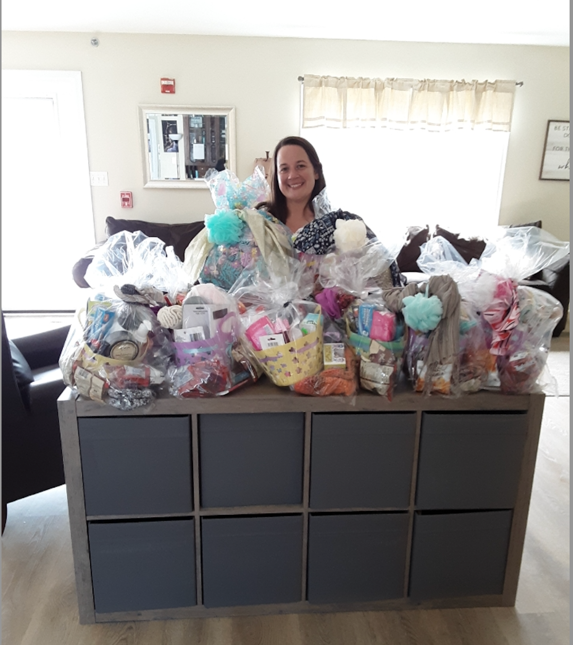 Woman smiles, surrounded by gift baskets on a gray cube shelf in a room with a window and a door.