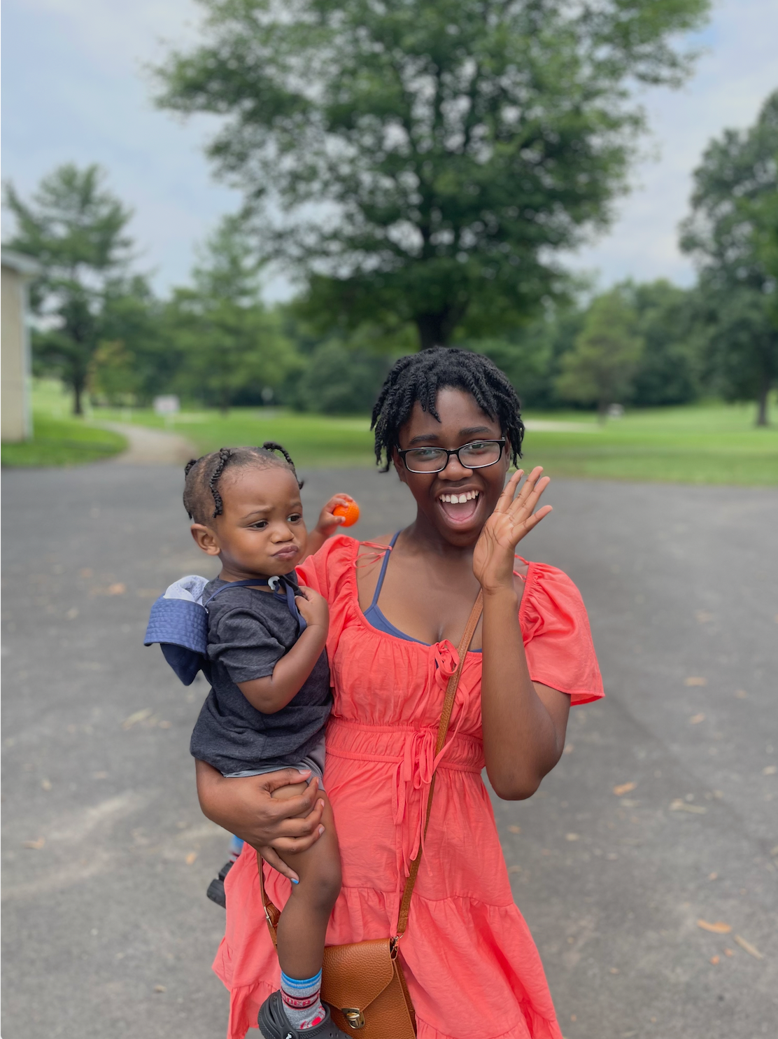 Woman in orange dress holding a child outdoors, smiling and waving. Child has a serious expression.