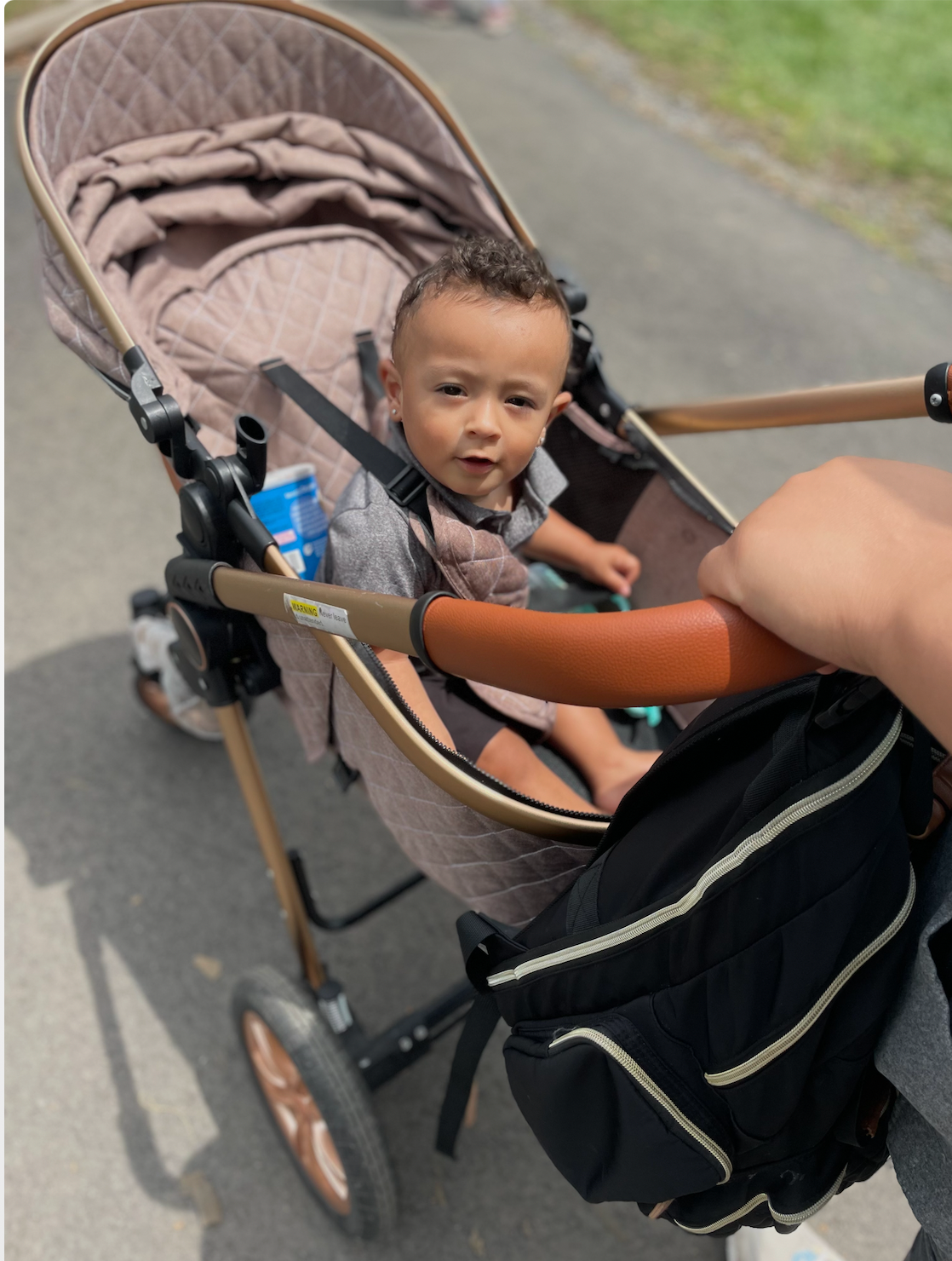 Child in stroller on a path, looking at the camera. Beige stroller with a brown handle and a black bag.