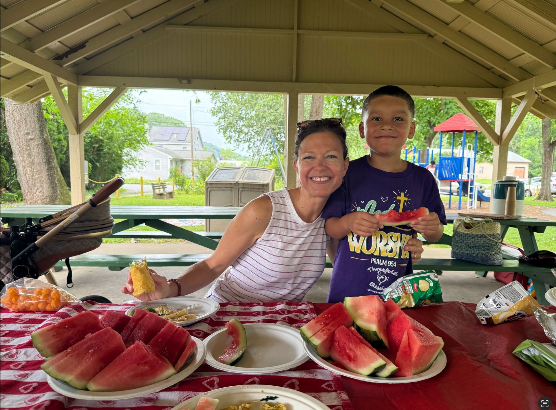 Woman and child smiling at picnic table with watermelon, snacks, and a playground in the background.