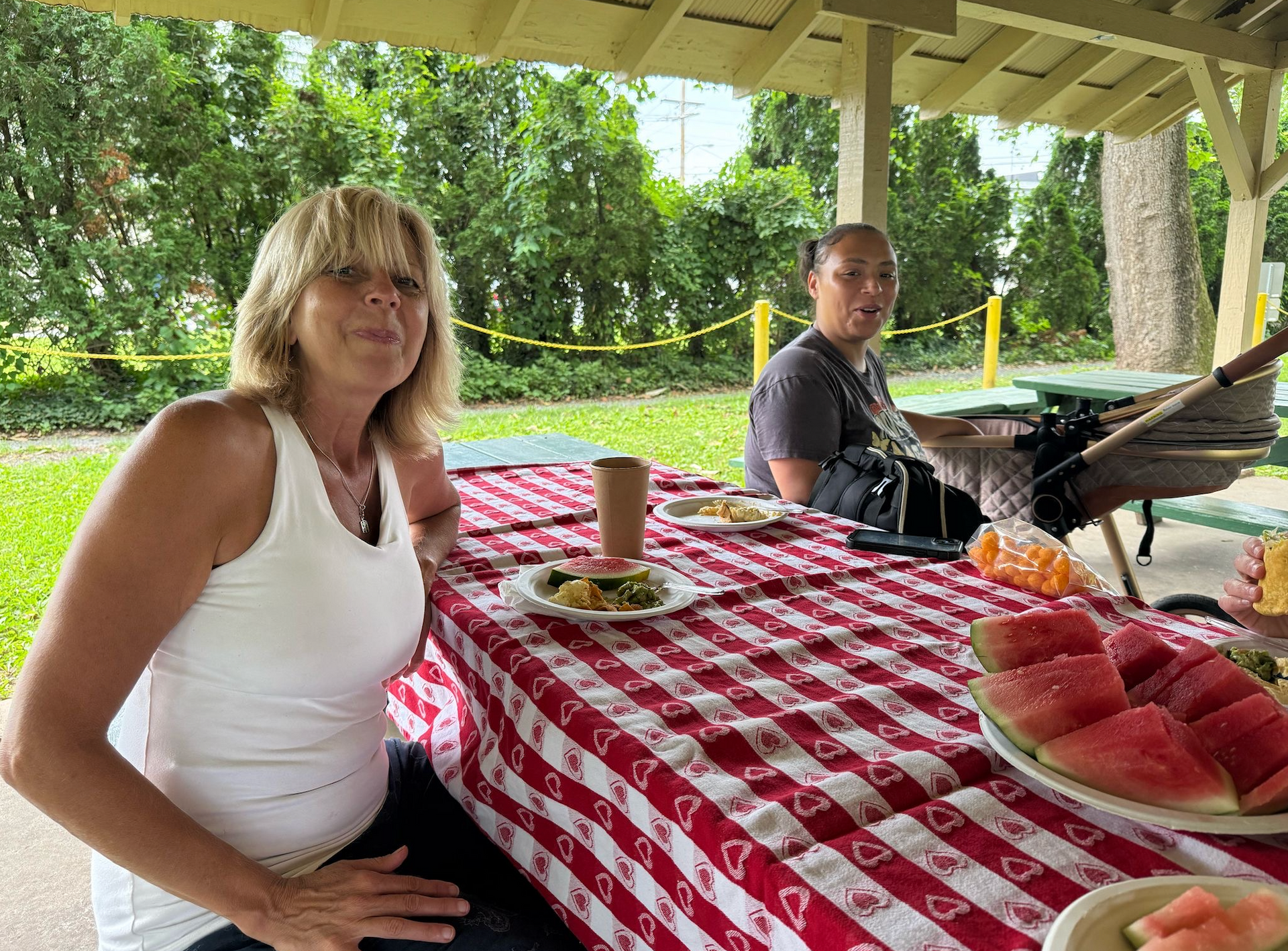 Two people at a picnic table with watermelon, food, and drinks in a park setting.
