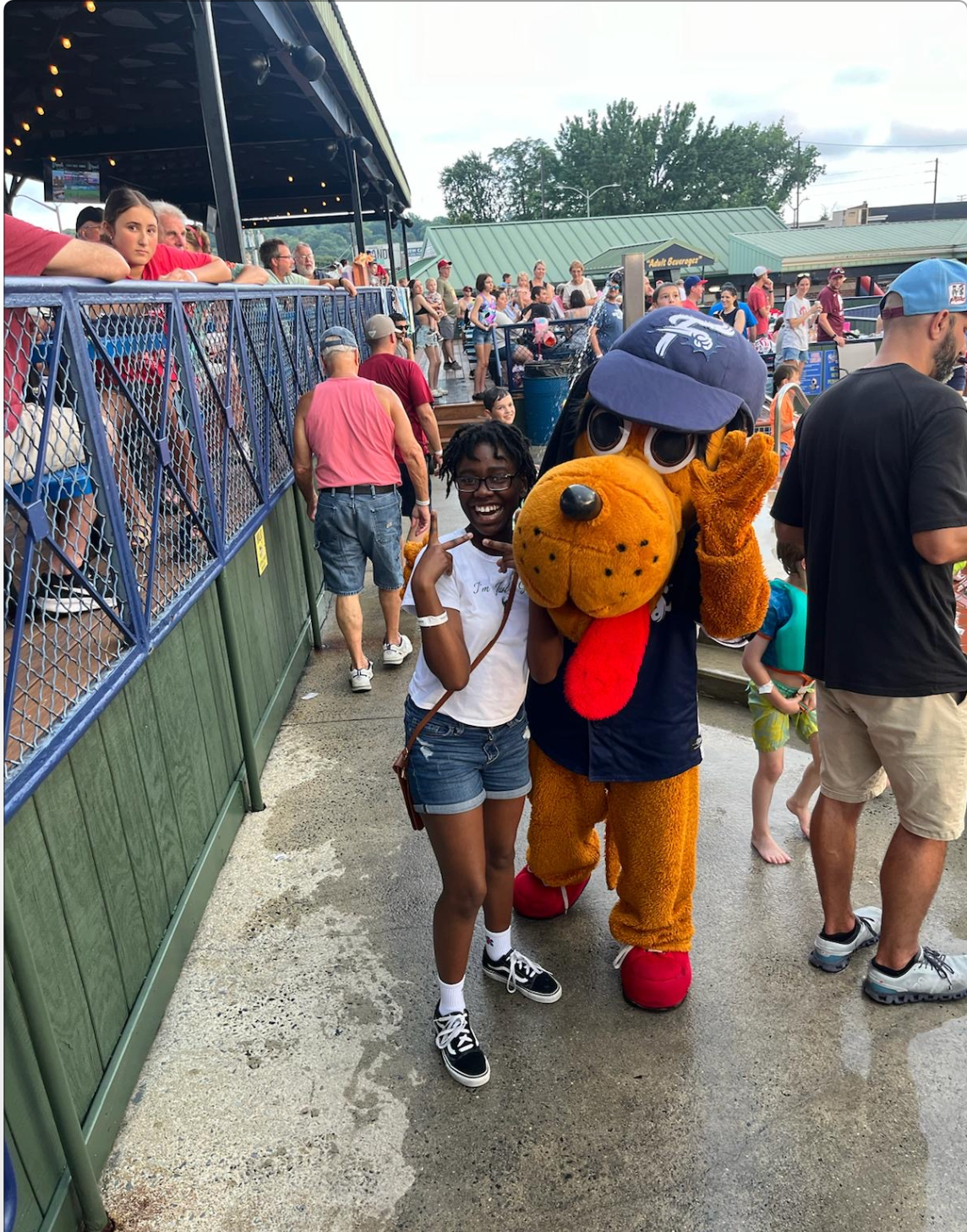 A young person smiles, making a peace sign, with a mascot wearing a baseball cap at an outdoor event.