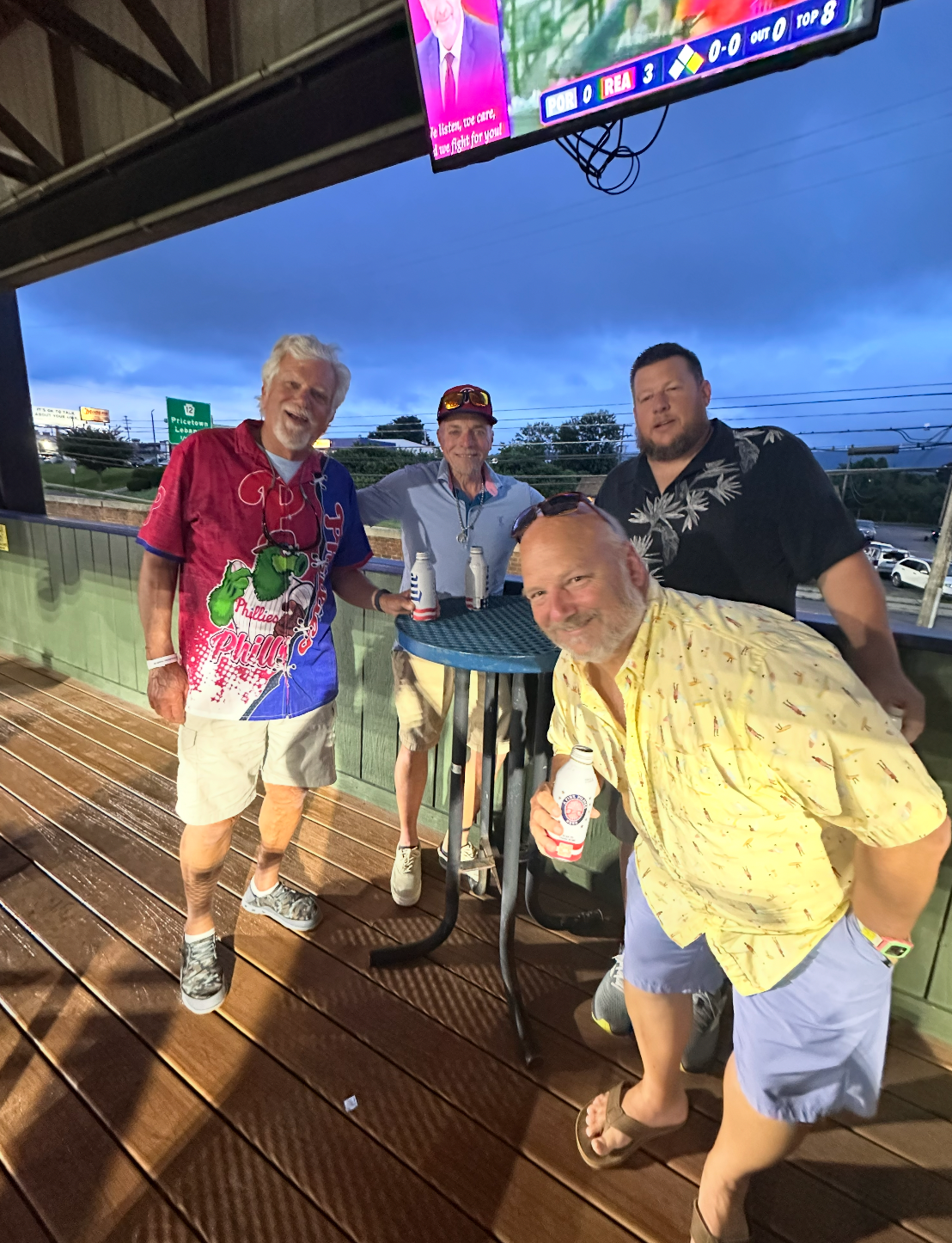 Four men smiling, holding drinks, on a wooden deck under an awning, watching TV.