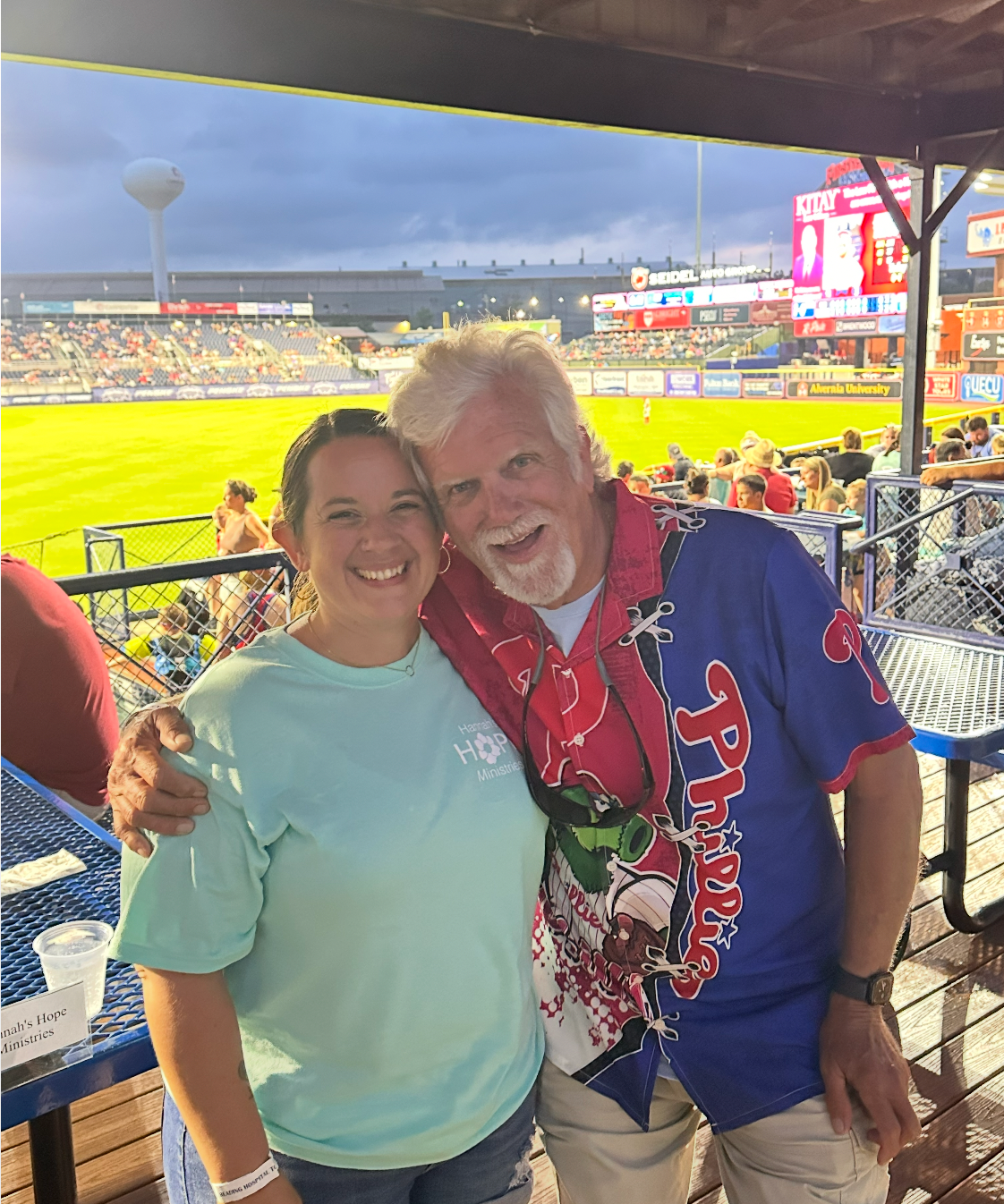 A smiling woman and man with his arm around her pose at a baseball game.