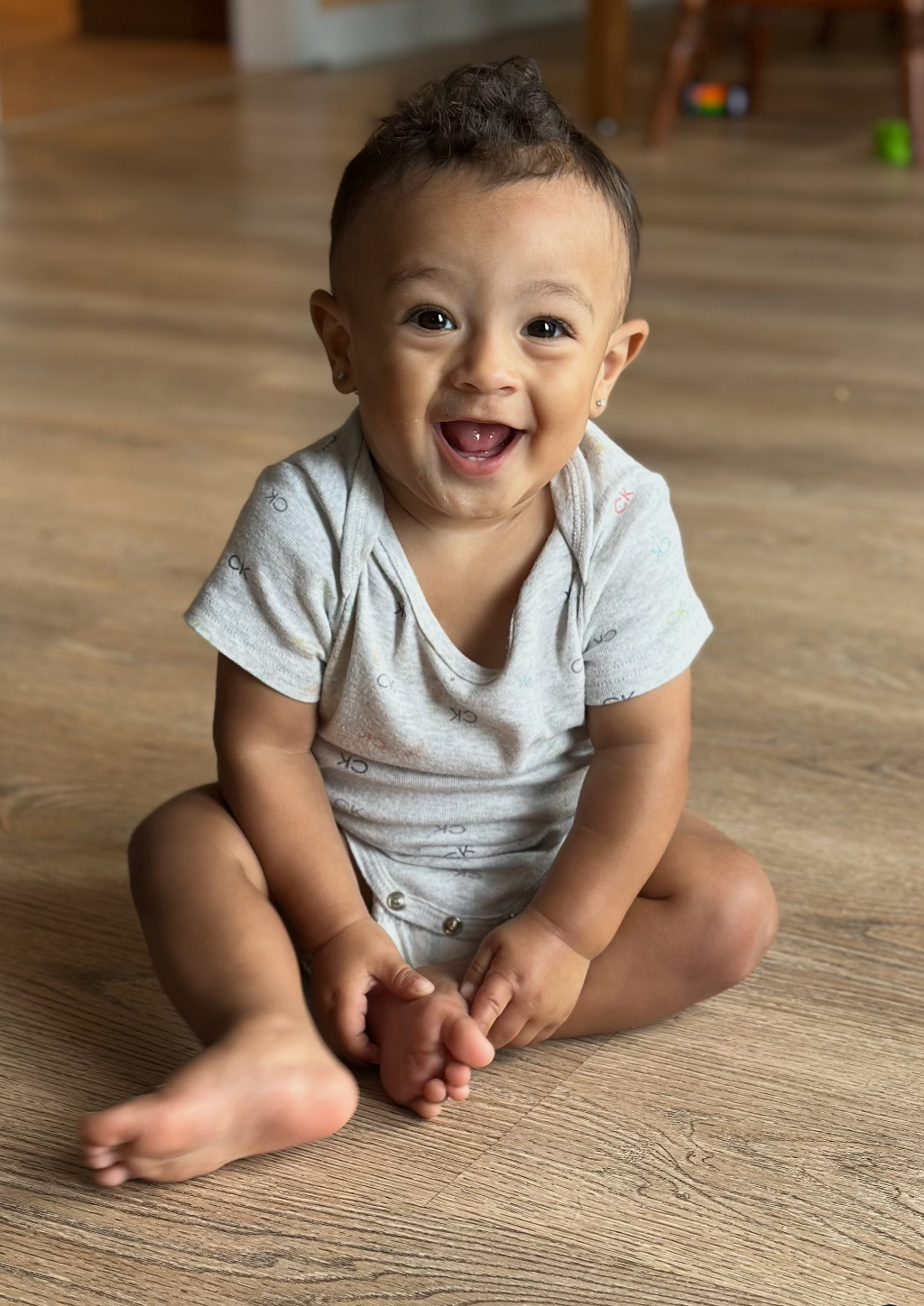 Happy baby sitting on the floor, smiling, wearing a gray onesie.