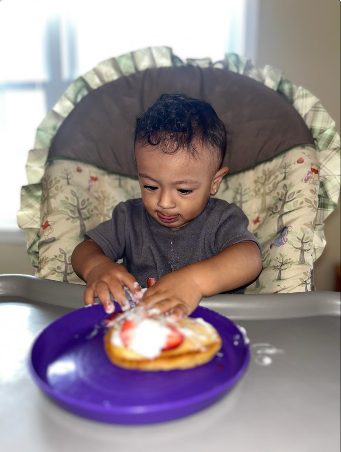 Child in high chair eating from a purple plate, food on hands and plate.