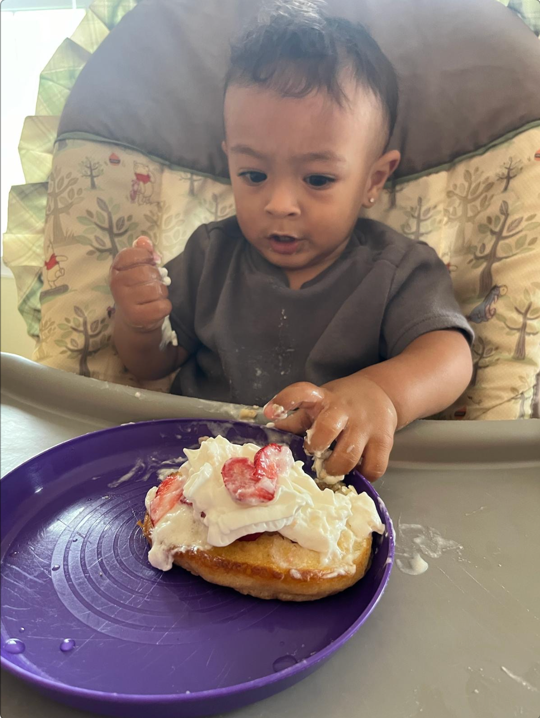 Baby in a high chair eating a pancake with whipped cream and strawberries, messy face and hands.